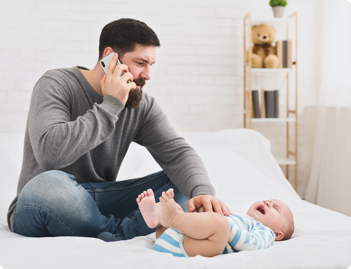 an image of a man sitting on a bed with his crying baby in front of him as he is talking on the phone with a concerned look on his face