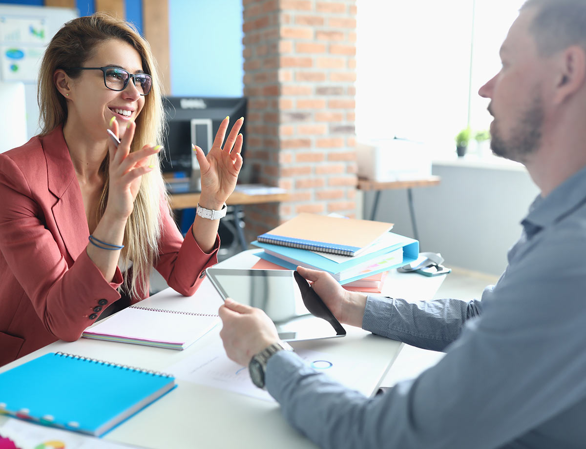 a man and a woman in business attire sitting across a desk from one another having a meeting