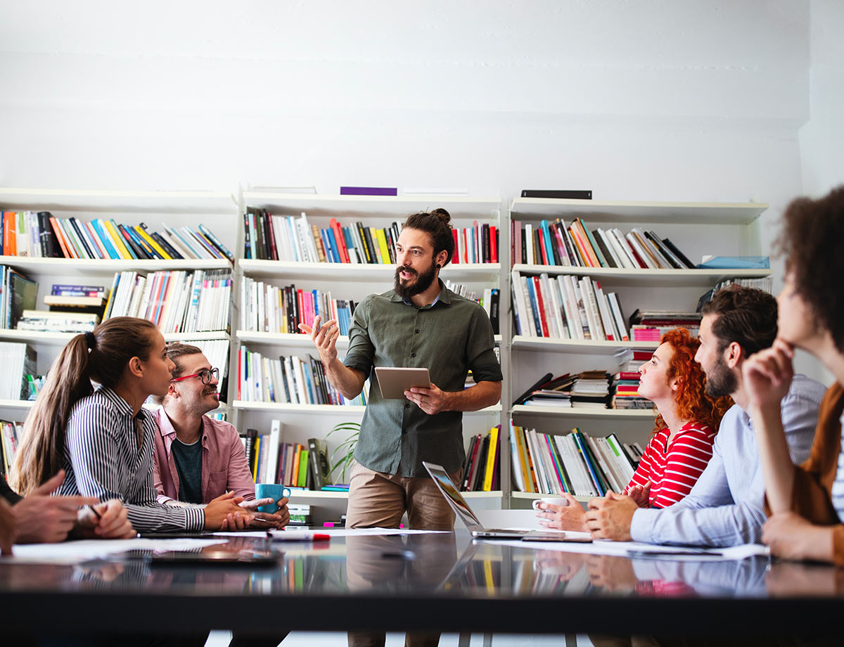 a man in a green shirt giving a presentation to a group of people sitting around a table in room lined with with book shelves