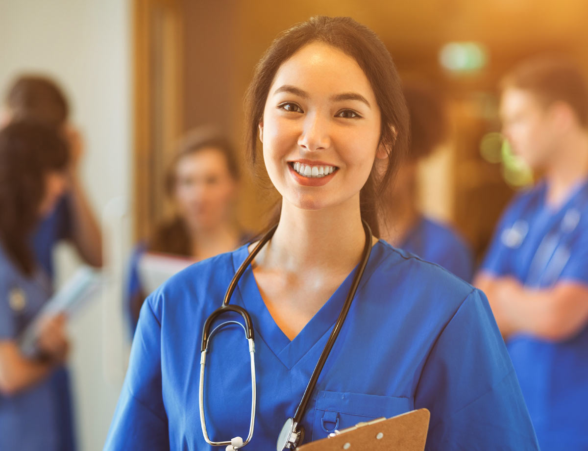 an image of a young female health care professional in blue scrubs smiling with people blurry in the background behind her