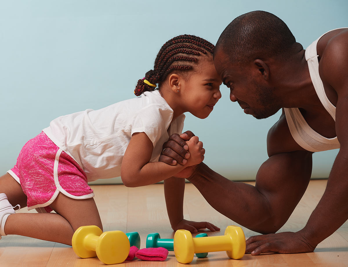 a little girl and her dad playfully arm wrestling on the floor with small fitness weights in front of them